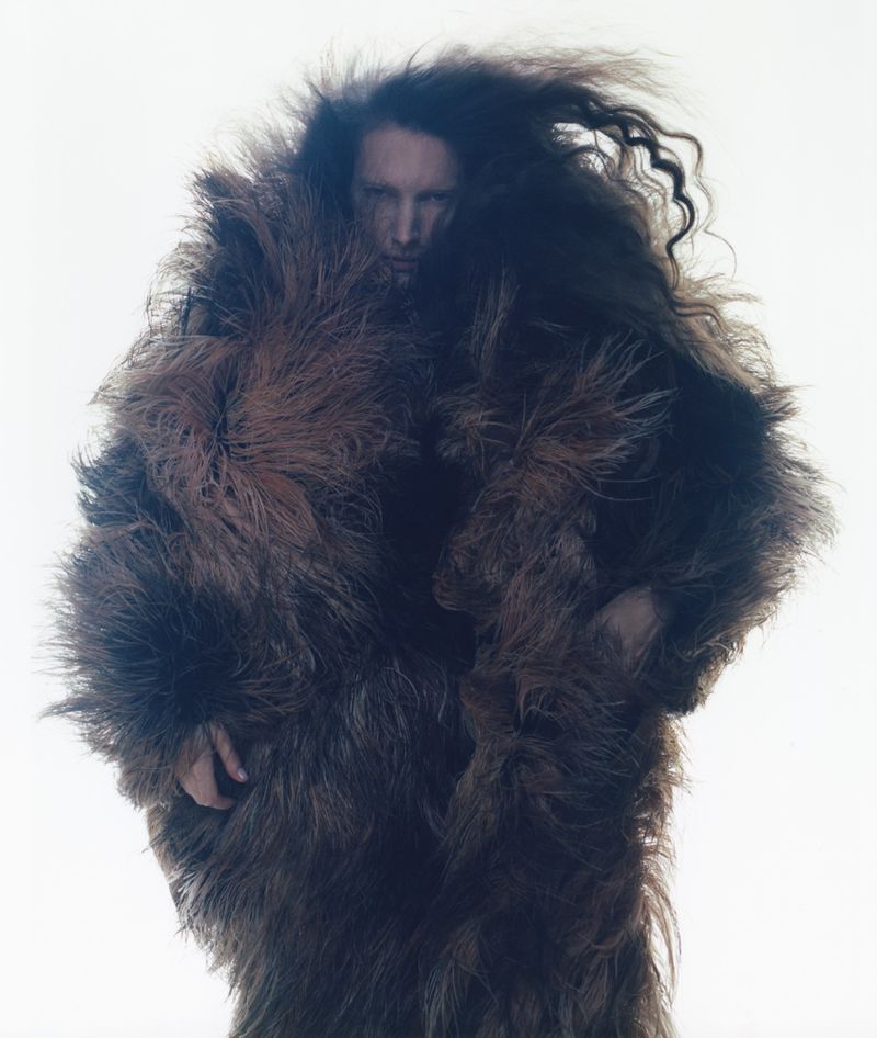 Adult stands indoors shrouded in a shaggy brown fur coat and flowing hair, set against a bright white background in a fashion editorial shot.
