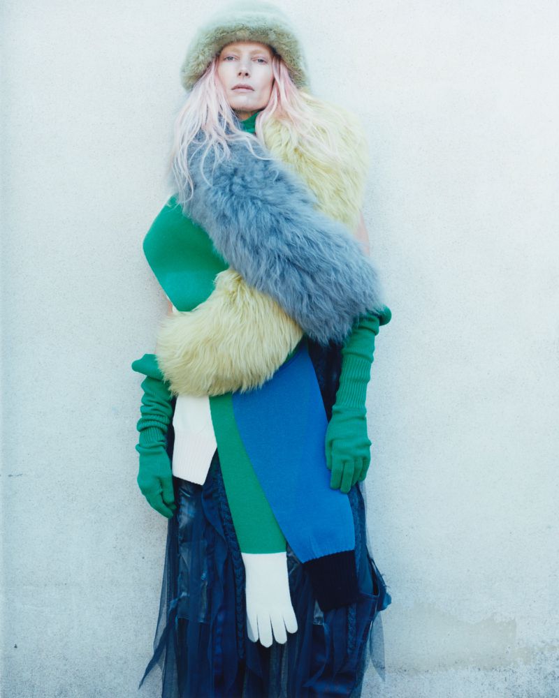 A fair-skinned woman with pink hair wearing a faux fur hat and scarf in blue, green, and yellow stands against a solid off-white wall.