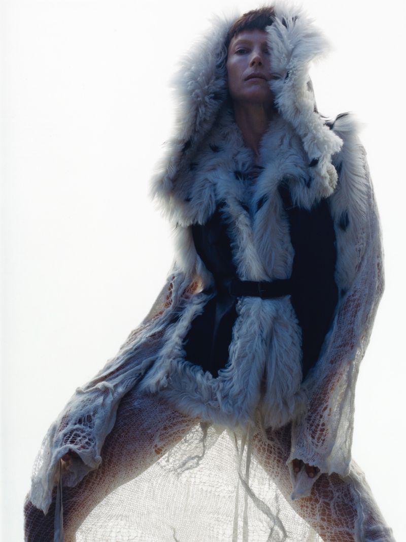 A woman models a dramatic fur-trimmed coat and matching pants against a stark white backdrop, blending high fashion with textural elements.