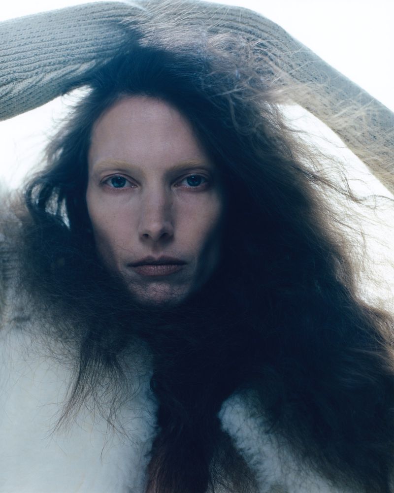 Close-up portrait of a woman with dark wavy hair, wearing a white furry coat and a knit sweater, set against a bright neutral background.