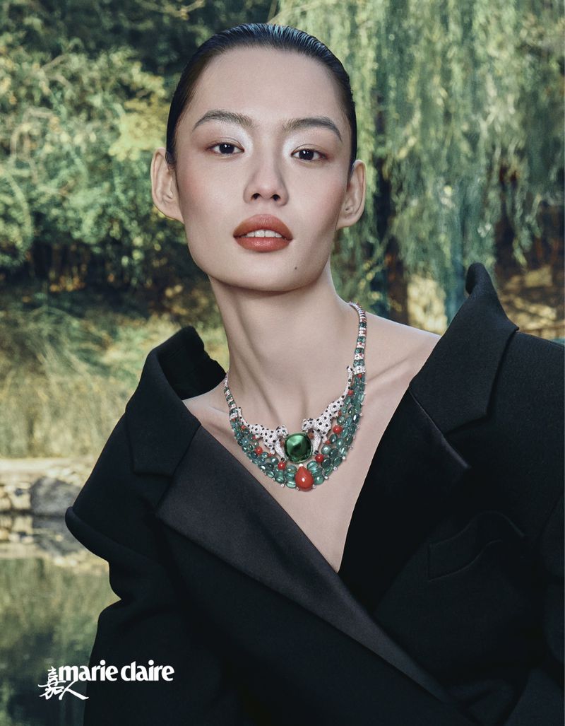 Close-up of an Asian woman wearing an ornate necklace and a black blazer, set against a blurred background of trees and water.