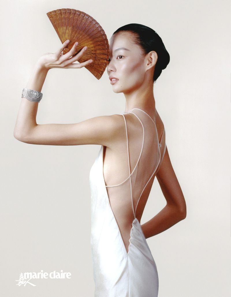 Elegant Asian woman poses in a white dress holding a fan, showcasing a Marie Claire fashion editorial against a pale background.