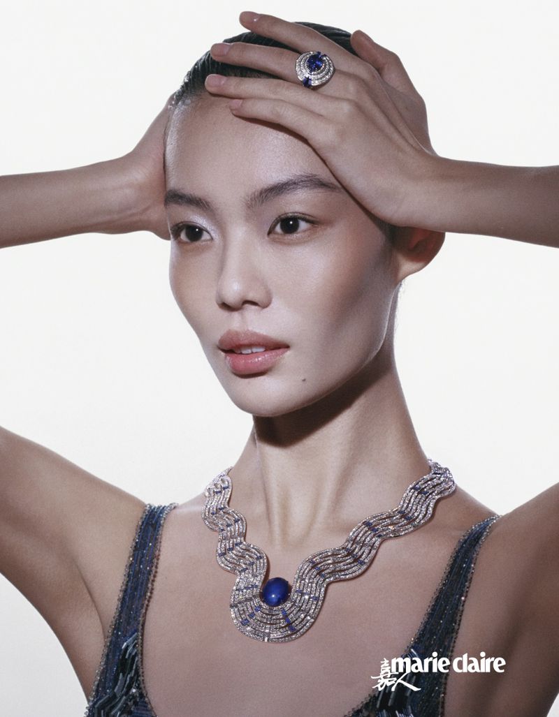 Elegant Asian woman with hands atop her head, showcasing jewelry: a necklace and large ring with a vibrant blue stone, against a bright background.