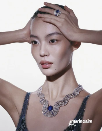 Elegant Asian woman with hands atop her head, showcasing jewelry: a necklace and large ring with a vibrant blue stone, against a bright background.