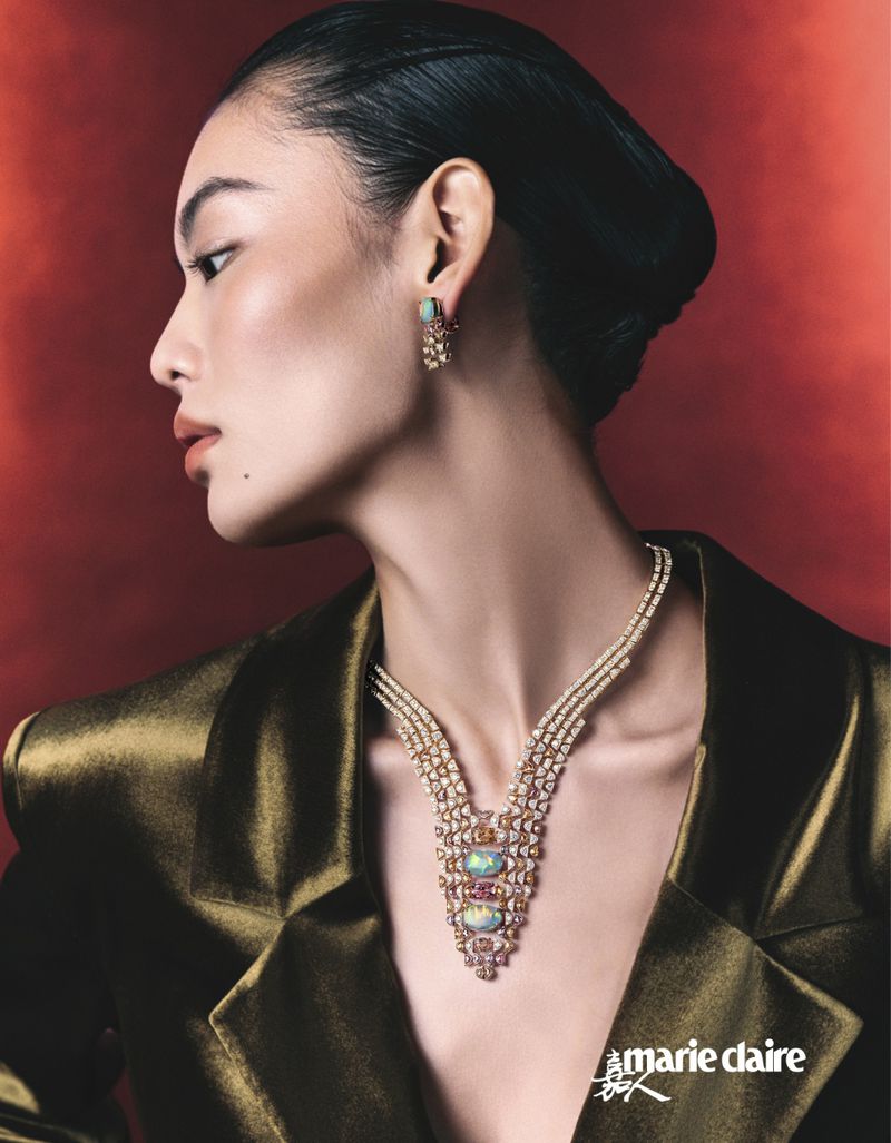 Elegant woman in a gold jacket wearing a jeweled necklace and earrings against a red backdrop in a beauty and fashion shot.