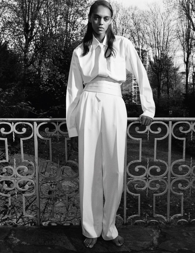 Elegant, barefoot adult posing in a matching white shirt and wide-leg pants near an ornate iron railing and a lush background of leafy trees.