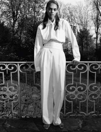 Elegant, barefoot adult posing in a matching white shirt and wide-leg pants near an ornate iron railing and a lush background of leafy trees.