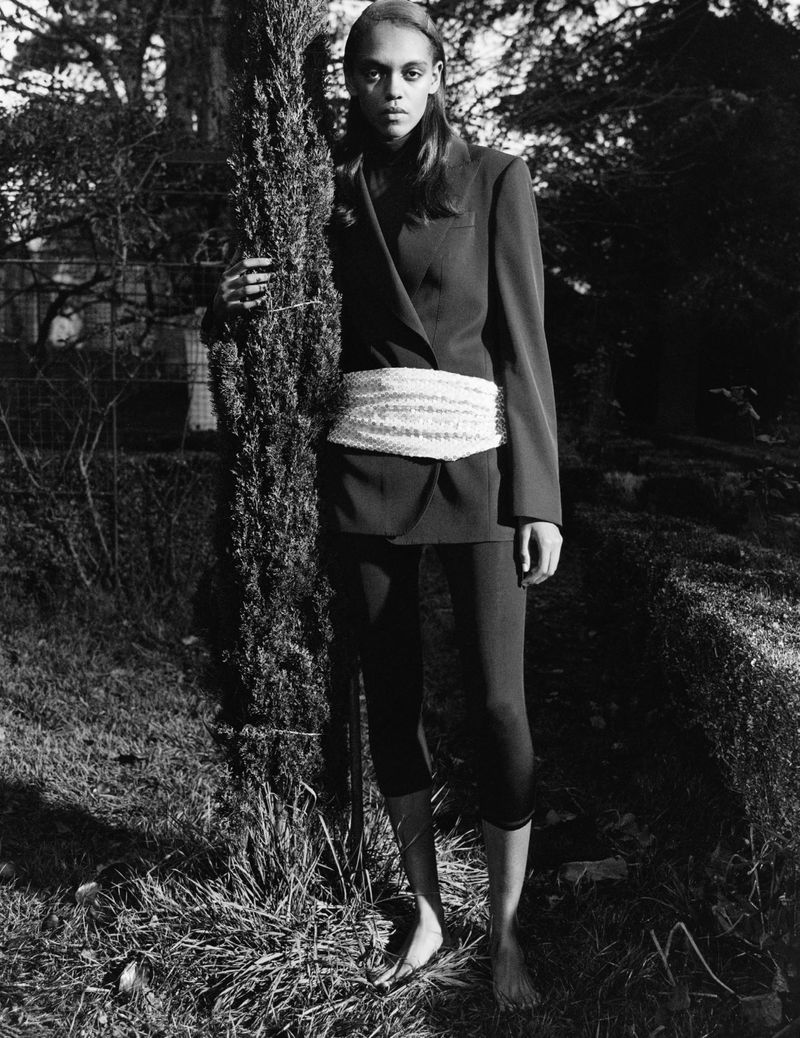 Fashionable adult woman poses in a black blazer and leggings near a coniferous tree in an outdoor wooded environment.