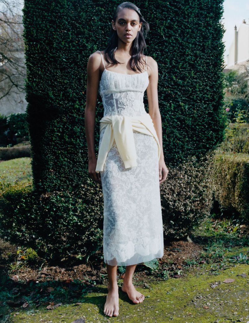 Woman in an embellished white dress with spaghetti straps poses barefoot outdoors in a grassy, garden-like setting with green hedges.