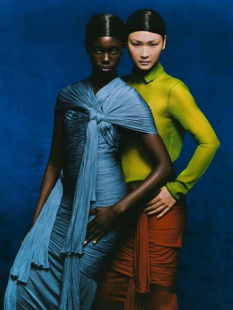 Two fashion models pose in draped dresses against a deep blue backdrop, showcasing a blend of textures and a modern, elegant aesthetic.