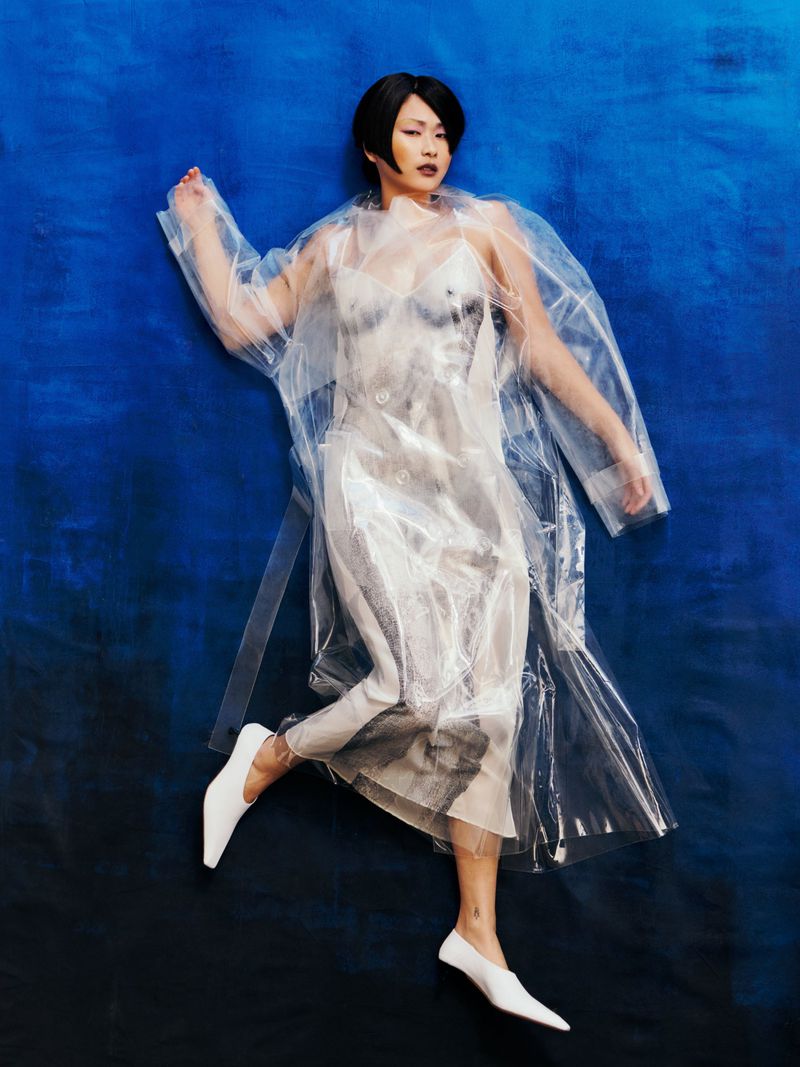 Overhead shot of a fashion model with short black hair lying on a blue background, wearing a sheer dress and clear plastic coat.