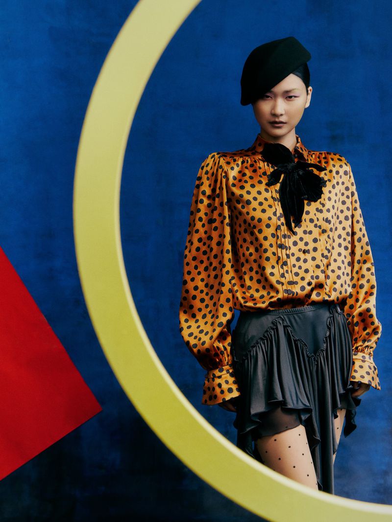 A stylish Asian woman poses against an abstract blue backdrop with yellow geometric shapes, wearing a polka dot blouse and black beret.