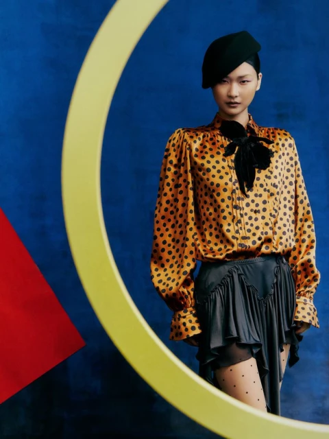 A stylish Asian woman poses against an abstract blue backdrop with yellow geometric shapes, wearing a polka dot blouse and black beret.