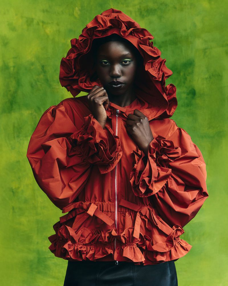 Fashion portrait of a Black woman wearing a ruffled red jacket with a hood, against a green textured background in a studio shot.