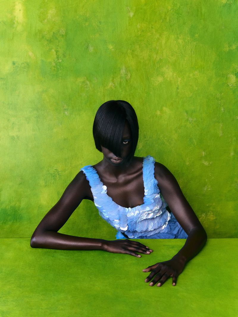 A dark-skinned model with a short black bob and side-swept bangs wearing a light blue sequined top against a textured lime green wall.