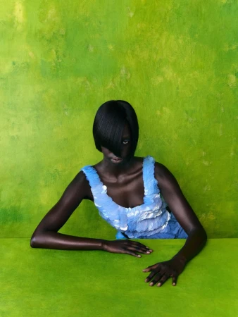 A dark-skinned model with a short black bob and side-swept bangs wearing a light blue sequined top against a textured lime green wall.