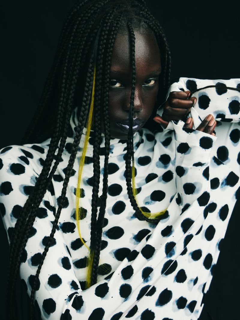 Close-up studio shot of a dark-skinned woman with box braids and yellow streaks wearing a black and white polka dot top.