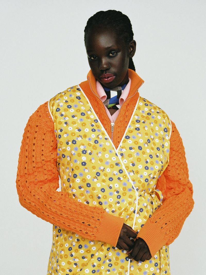 Black model wearing an orange patterned floral dress and knitted cardigan against a plain white background.