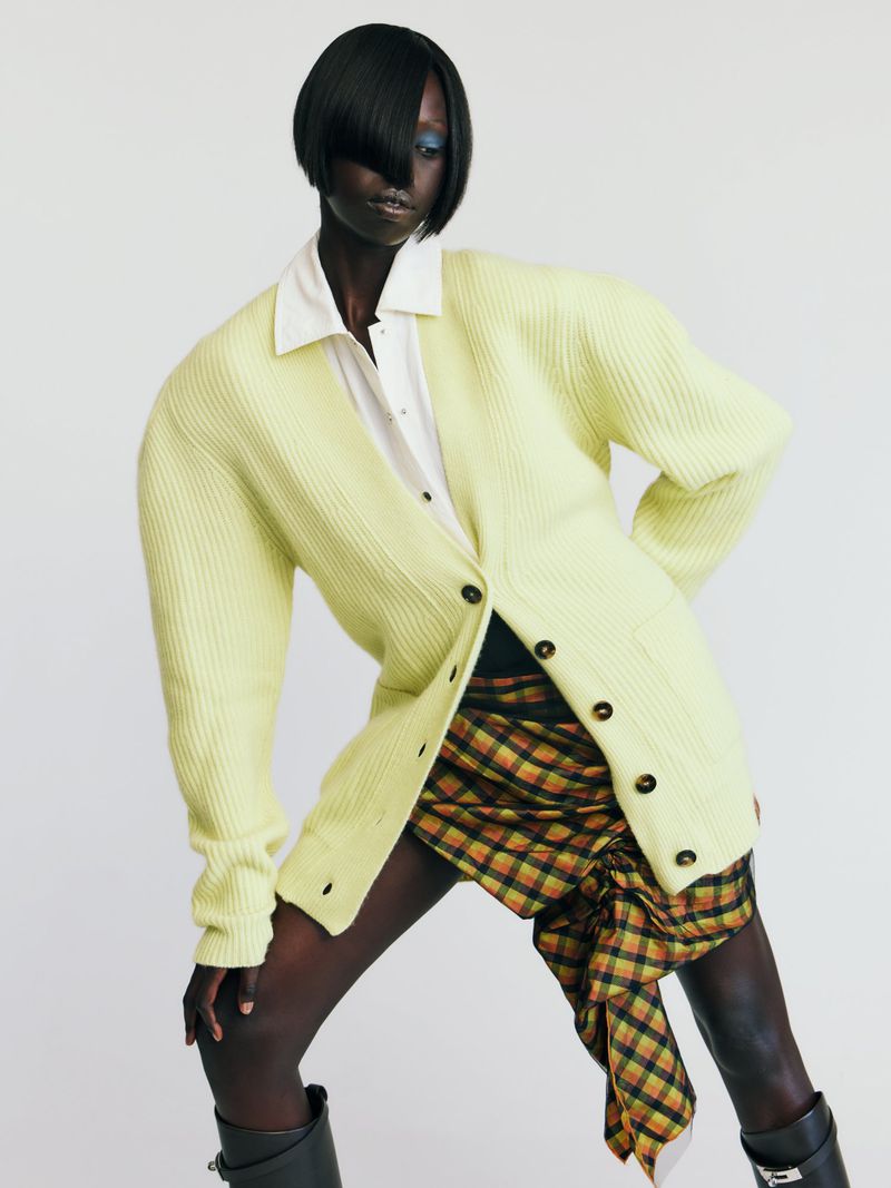 An adult woman poses in a white collared shirt, yellow cardigan, plaid skirt, and black boots against a white background in a fashion shot.