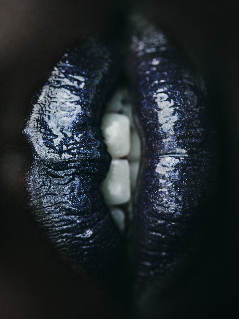 Extreme close-up of dark, glossy lips parted to reveal white teeth in a beauty or fashion image with high contrast and bold color.