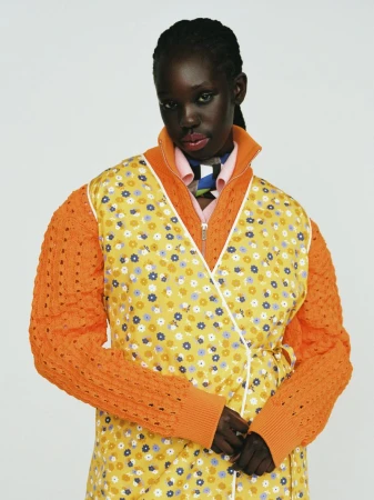 Black model wearing an orange patterned floral dress and knitted cardigan against a plain white background.