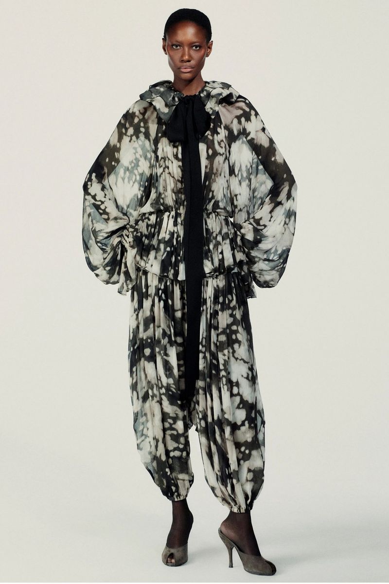 Editorial fashion shot of an African woman in a hooded black and white jumpsuit with heeled shoes, against a neutral backdrop.