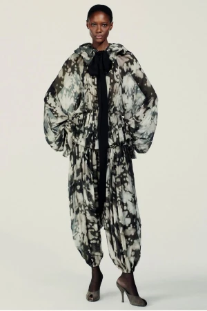 Model wearing a patterned voluminous hooded outfit with a black ribbon tie against a plain backdrop.