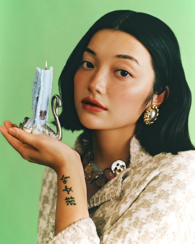 Portrait of an Asian woman holding a lit candle, wearing pearl earrings and a tweed jacket against a light green background.