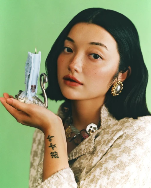 Portrait of an Asian woman holding a lit candle, wearing pearl earrings and a tweed jacket against a light green background.