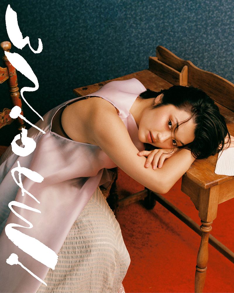 Young woman with short dark hair rests head on table wearing light pink top. Shot of face, shoulders, and desk indoors.