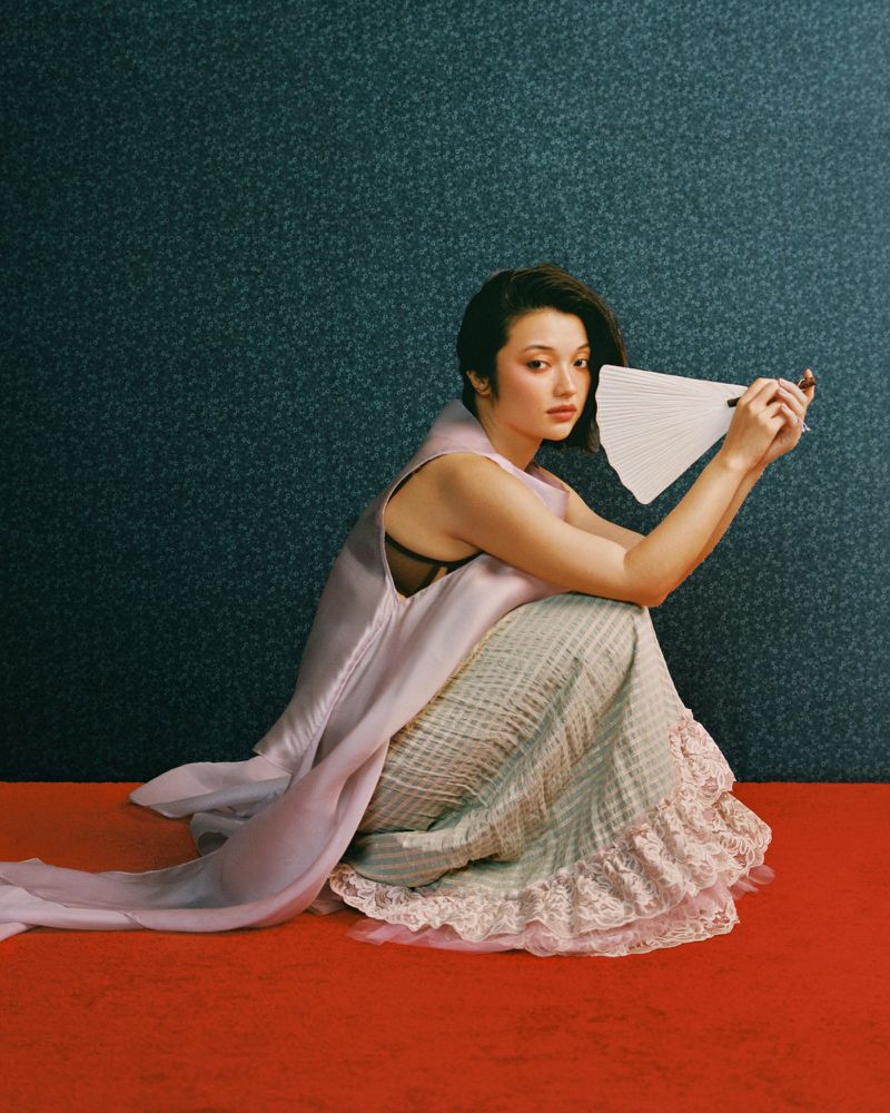 Young woman with short hair, kneeling, and holding a white fan. She wears a pastel gown against a dark, patterned backdrop.