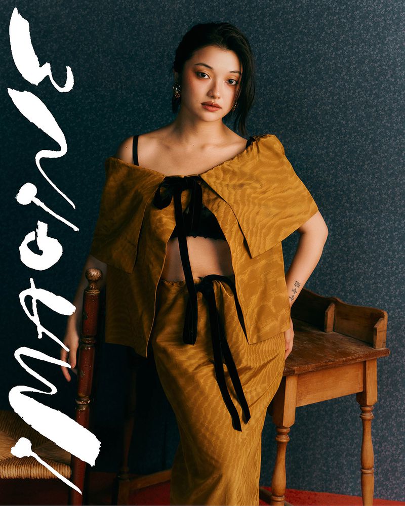 Woman poses indoors wearing a mustard-colored outfit with black ribbon details, complemented by antique wooden chair and table.
