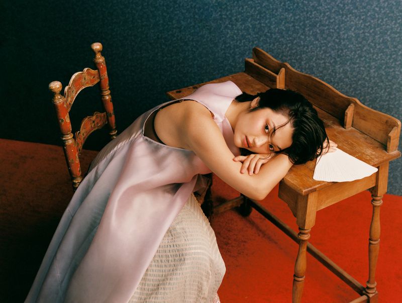 A young adult woman in a lilac dress rests her arms on a wooden desk with a solemn facial expression in a warmly lit indoor setting.