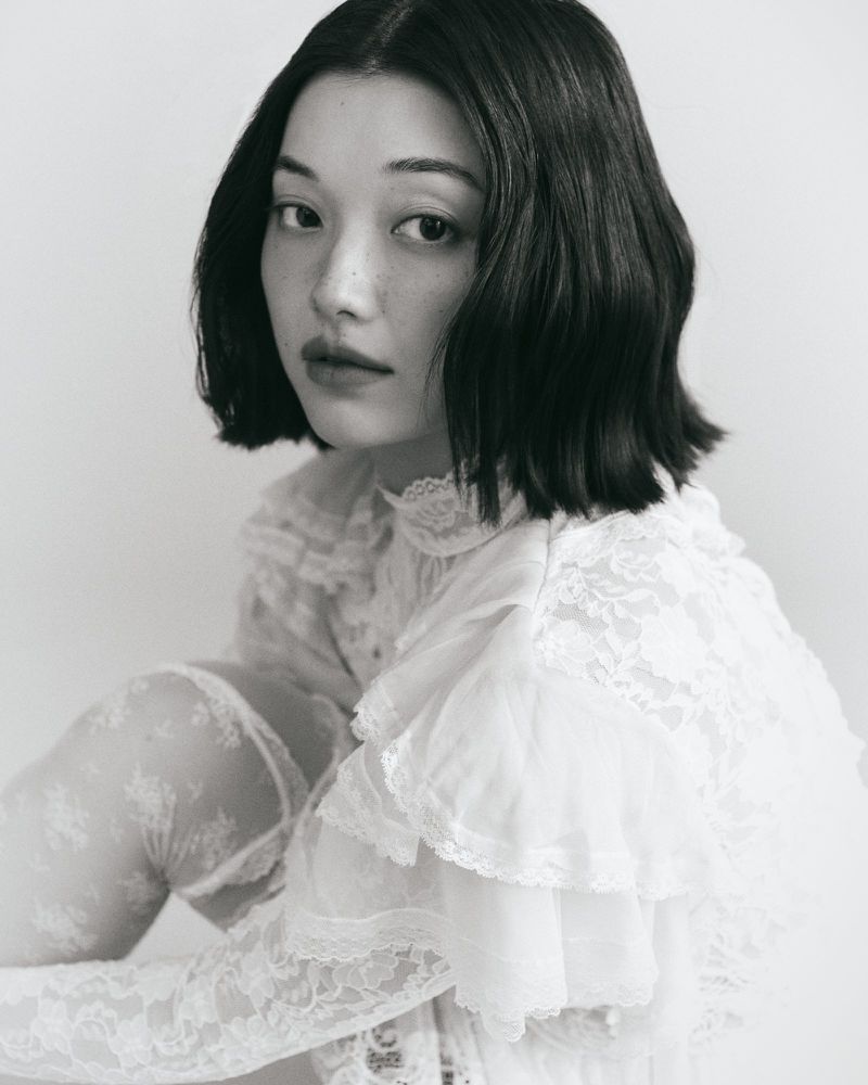 Black and white portrait of an Asian woman with dark hair in a white lace top, gazing intently at the camera, soft lighting, and pale background.