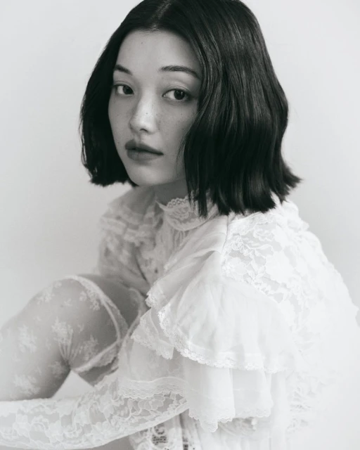 Black and white portrait of an Asian woman with dark hair in a white lace top, gazing intently at the camera, soft lighting, and pale background.