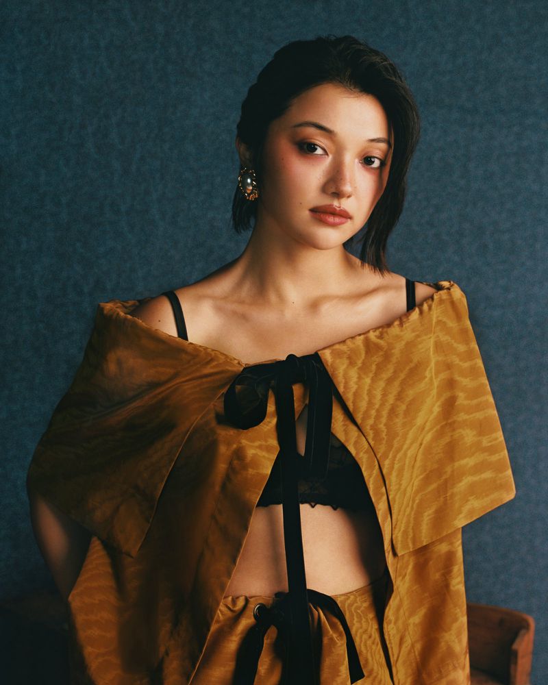 A woman with dark hair poses in a studio with fair skin and golden brown dress with black bows, against a textured blue backdrop.