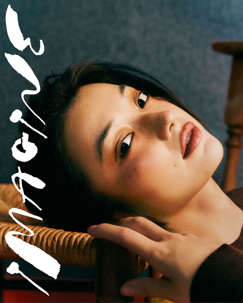 Portrait of an Asian woman with short black hair, fair skin, and freckles, lying on a wooden chair with white text overlay on a gray background.