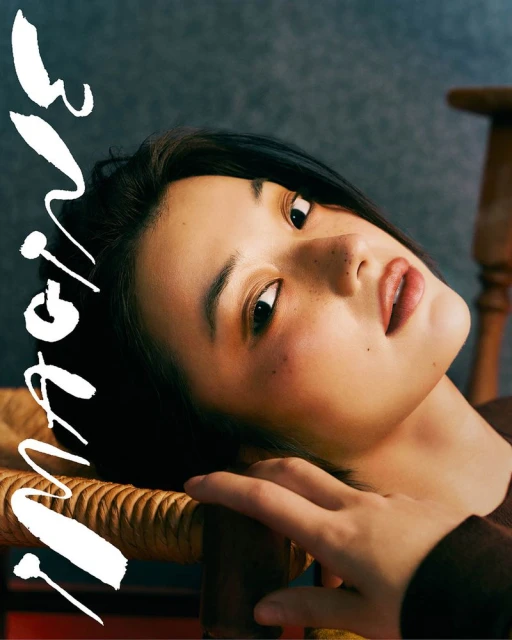 Portrait of an Asian woman with short black hair, fair skin, and freckles, lying on a wooden chair with white text overlay on a gray background.