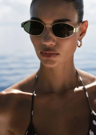 Adult woman wearing stylish sunglasses and a polka dot bikini top poses against a backdrop of the ocean and sky on a sunny day.