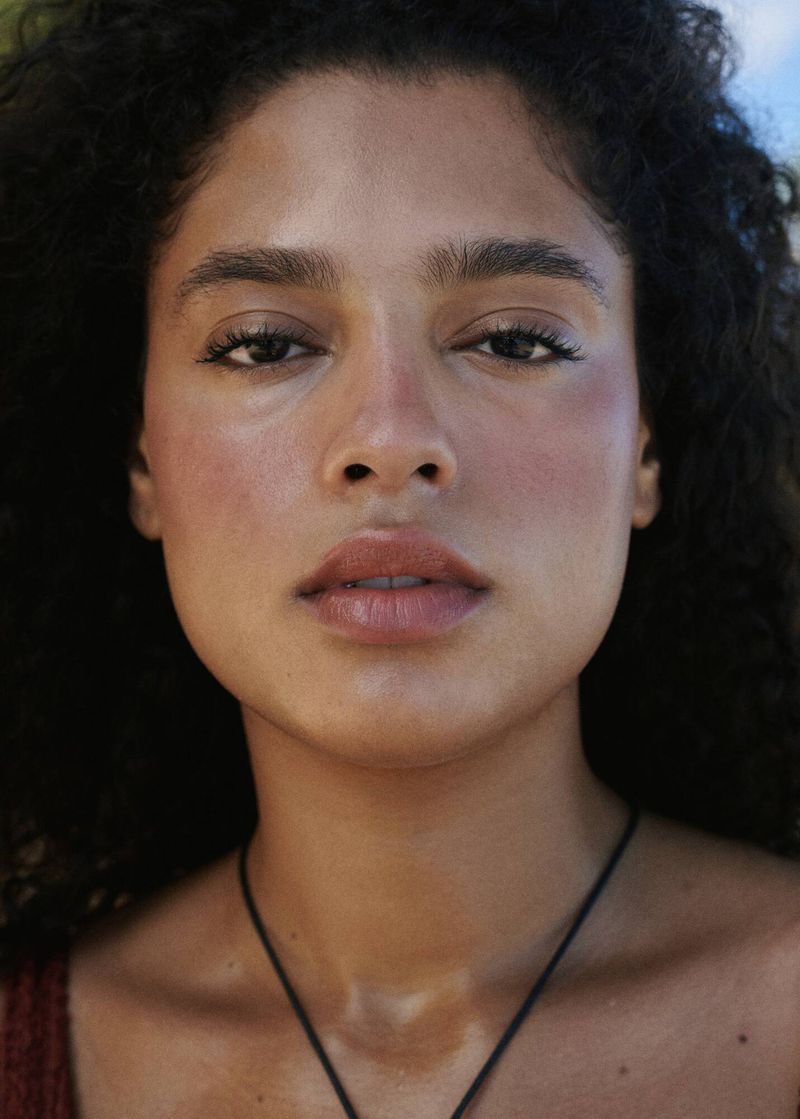 Close-up portrait of an adult female with curly hair, brown eyes, and a necklace outdoors, illuminated by natural light on her face.