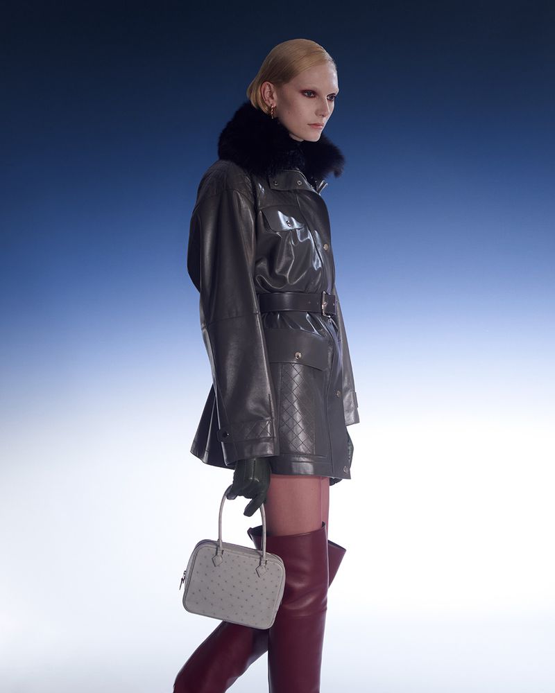 Fashion model wearing a fur-trimmed leather coat, thigh-high boots, and carrying a structured bag against a blue gradient background.