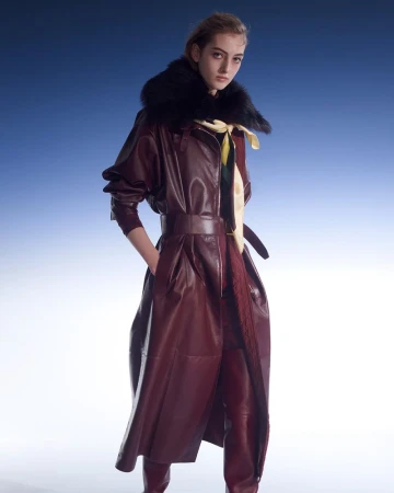 A single young woman with light skin, wearing a burgundy leather trench coat with a black fur collar, stands against a blue background.