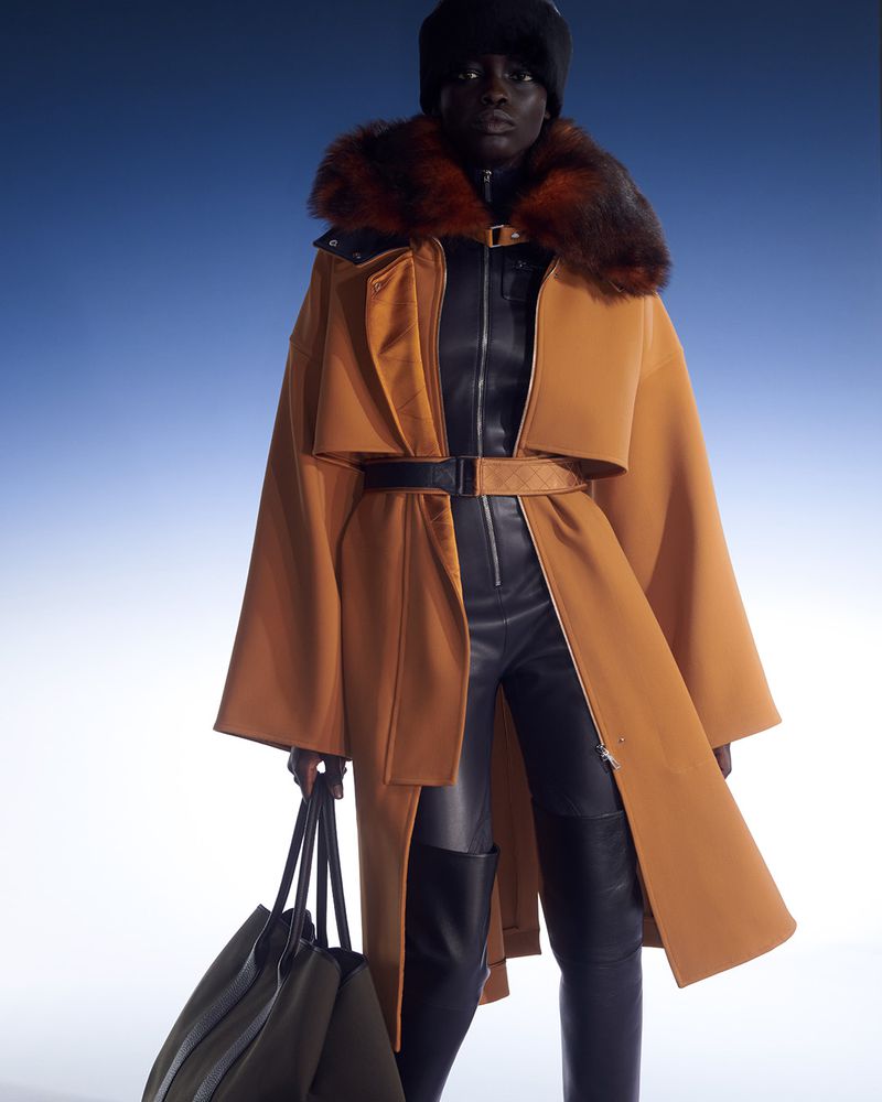 Fashion shot of a Black woman wearing a long, tan coat with fur collar, black bodysuit, heeled boots, and carrying a tote bag.