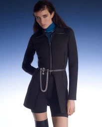 Model wearing a black zip jacket with a chain belt over shorts and blue turtleneck against a gradient blue background