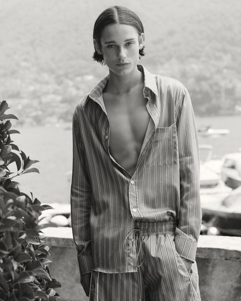 Monochromatic medium shot of a young adult male wearing a striped pajama set, posing outdoors against a blurred background of water and greenery.