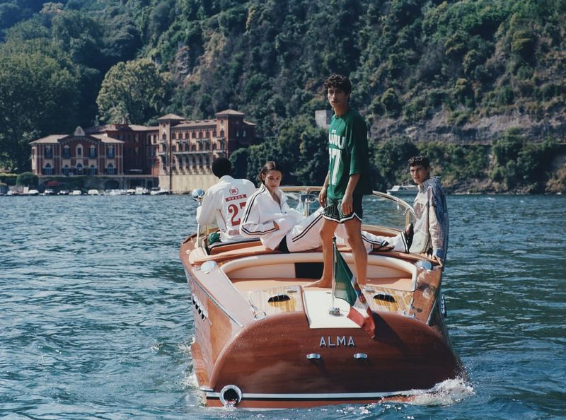 Four young adults aboard a wooden speedboat cruising on a lake with lush green mountains and a villa in the background, on a sunny day.