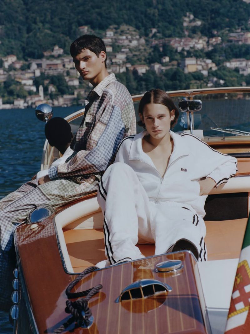 Two men sit on a wooden boat in casual outfits with a lake and town in the background on a sunny day, fashion editorial style.