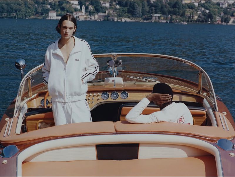 Two people in matching tracksuits sit in a wooden boat on a lake surrounded by mountains in daytime, enjoying the scenic views.