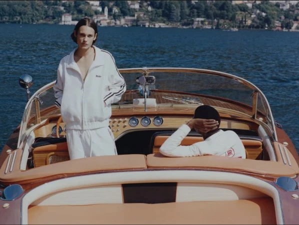Two people in matching tracksuits sit in a wooden boat on a lake surrounded by mountains in daytime, enjoying the scenic views.