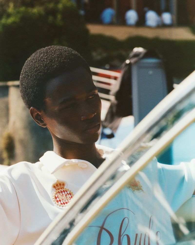 Man in light polo shirt stands outdoors near water, partially obscured by a reflective glass surface on a sunny day. 